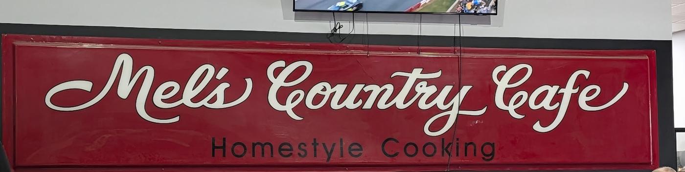 The original hand-painted board reading Mel's Country Cafe, Homestyle Cooking, in white script on red, mounted on the dining room wall.